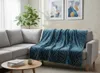 A blue throw blanket with a geometric pattern is draped over a light gray sectional sofa in a living room setting. Two abstract art pieces hang on the wall behind the sofa, and a potted plant is visible to the left. A small round coffee table with books an Podgląd zdjęcia produktu