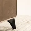 A close-up view of the leg of a brown upholstered bed frame resting on a cream-colored rug. Produktbild-Vorschau
