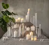 A collection of LED candles of various sizes and shapes arranged on a tiered display with a large green plant in the background. Produktbild-Vorschau