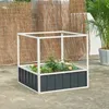 A raised garden bed with a white frame and dark gray corrugated sides, containing several small plants and flowers. It is placed on a concrete surface, with a woven bamboo-like screen and a potted palm tree in the background. Produktbild-Vorschau