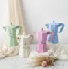 Four colorful Moka pots are arranged on a marble surface with draped fabric and dried flowers. Produktbild-Vorschau