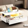A modern living room scene featuring a white coffee table with drawers and a lifted section, a beige sofa, and some decorative items. Produktbild-Vorschau