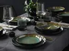 A beautifully set dining table with a dark tablecloth, featuring a set of stoneware plates, bowls, glasses, and cutlery, with a decorative vase and plant in the background. Náhľad obrázku produktu