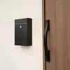 A black mailbox mounted on a white wall next to a wooden door with a black handle. Voir les images du produit