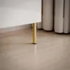 A close-up shot of the bottom of a white furniture piece with a gold leg resting on a light wood floor. Náhled obrázku produktu