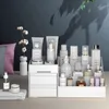 A vanity or makeup organizer filled with various cosmetic products, including bottles, jars, and tubes, displayed on a desk. A small vase with cotton stems is also present. Náhľad obrázku produktu