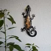 A decorative solar-powered gecko light is mounted on a textured wall, with a leafy plant in the foreground. Produktbild-Vorschau