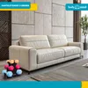 A cream-colored sofa is shown in a modern living room setting, with a rack of colorful dumbbells in the foreground. Náhľad obrázku produktu