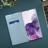 A light blue flip case for a Samsung Galaxy S20 phone is shown open, with a credit card in the case's slot and the phone displaying a colorful wallpaper. A keyboard and a potted plant are visible in the background. Anteprima dell’immagine prodotto