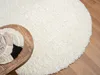 A close-up view of a round, white, shaggy rug on a wooden floor, with a small wooden table in the foreground holding coasters. Náhľad obrázku produktu