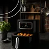 A black air fryer with its basket pulled out, filled with food, sits on a countertop next to a potted plant and a small bowl of salt. Produktbild-Vorschau