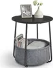 A round side table with a black top and gray fabric basket underneath, holding books and a blanket. A framed picture and a vase with flowers are on top. Produktbild-Vorschau