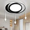 A modern LED ceiling light with a black and white ring design, shown mounted on a white ceiling. Arrows point from different parts of the light fixture to labels indicating materials. Náhled obrázku produktu