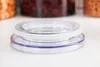 A close-up shot of a clear plastic jar lid with a white seal and blue trim, resting on a white surface. In the background, out of focus, are jars containing food items. Produktbild-Vorschau