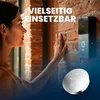 A woman is interacting with a smart doorbell mounted on a brick wall, with a coin cell battery in the foreground. Produktbild-Vorschau