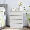 A white chest of drawers with four drawers, decorated with two photo frames and a vase with flowers on top, next to a bed with a grey headboard and a striped duvet. Náhľad obrázku produktu