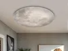 A ceiling light fixture resembling the moon is installed on a white ceiling in a room with gray walls. Artwork is visible on the wall. Náhled obrázku produktu