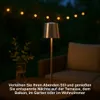 A table lamp is placed on a table next to a vase of greenery, with a couch and string lights in the background, suggesting an outdoor evening setting. Podgląd zdjęcia produktu