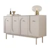 A light beige sideboard with four doors and gold handles, standing on wooden legs. It is decorated with a lamp, a bowl, and a diffuser. Produktbild-Vorschau