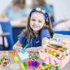 A young girl is smiling at the camera while doing crafts at a table. A pink craft box is open and filled with various craft supplies. Crafting materials are spread out on the table in front of her. Produktbild-Vorschau