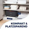 A dark gray appliance with a red trim is placed on a white shelf inside a cabinet. There are folded towels and storage bins on the shelves above and to the side. Text overlays indicate dimensions and marketing slogans. Produktbild-Vorschau