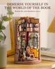A miniature 3D wooden book nook model of a study, filled with books, a ladder, a desk, and a chair, placed on a wooden surface with a blurred background of a plant and a book. Náhled obrázku produktu