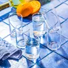 Six clear drinking glasses filled with water, arranged on a blue tiled surface, with oranges and a bottle in the background. Voir les images du produit