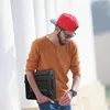 A man wearing a red baseball cap and sunglasses walks outdoors, holding a black laptop case. Produktbild-Vorschau