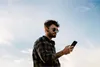 A man wearing sunglasses and a plaid shirt looks at his phone while standing outdoors against a blue sky. Produktbild-Vorschau