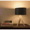 A table lamp with a black shade and a white tripod base sits on a dark wooden surface next to a stack of books. Voir les images du produit