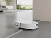 A white robot vacuum cleaner is cleaning a modern living room with large windows and furniture in the background. Náhled obrázku produktu