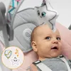 A baby is lying in a pink and gray baby bouncer with a toy star hanging above its head. There is a circular graphic in the bottom left corner with illustrations of a hot air balloon, musical notes, and a star. Produktbild-Vorschau