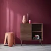 A wooden sideboard with a slatted sliding door, a vase, and candles on top, next to a pink stool against a dark pink wall. Anteprima dell’immagine prodotto