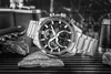 A black and silver chronograph watch is displayed on a textured surface with rocks and other objects in the background. Voir les images du produit