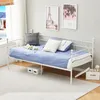 A white metal daybed with blue bedding and pillows in a room with a window and artwork on the wall. Náhled obrázku produktu