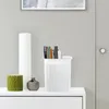 A white storage bin with books inside, a green candle, and a small gray vase on a white surface. Produktbild-Vorschau