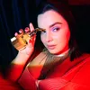 A woman holds a gold perfume bottle up to her eye, with dramatic red and blue lighting. Produktbild-Vorschau