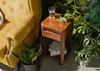 A wooden side table next to a yellow armchair, surrounded by lush green plants. Anteprima dell’immagine prodotto