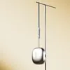 A silver-colored wireless earbud case is hanging from a metal stand against a light yellow background. Náhled obrázku produktu