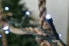 A close-up of a white LED light on a string of Christmas lights, with a blurred background of a Christmas tree and rope. Produktbild-Vorschau