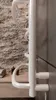 A close-up view of a white electric towel warmer mounted on a gray tiled wall, with a stone wall and wooden counter visible in the background. Náhľad obrázku produktu