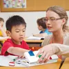 A teacher is helping a young boy with a Lego pencil case in a classroom. Produktbild-Vorschau