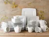 A collection of white porcelain dinnerware, including plates, bowls, cups, and a sugar bowl, arranged on a wooden surface with sprigs of yellow flowers. Náhľad obrázku produktu