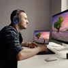 A man wearing headphones is working on a laptop and a tablet, with a large monitor in the background. Produktbild-Vorschau