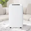 A white dehumidifier is placed on a fluffy white rug in a living room setting, with a couch and plant in the background. Náhled obrázku produktu
