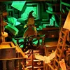 A miniature diorama of a witch's study, complete with a witch figure, spider, owl, books, and furniture, illuminated by green light. Voir les images du produit