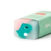 A close-up view of a teal dinosaur-shaped eraser with pink accents, partially emerging from a light blue translucent material, set against a white background. The eraser is inside a green cardboard box with text and leaf motifs. Produktbild-Vorschau