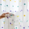 A hand holding a sheer curtain with pom-pom decorations. Produktbild-Vorschau