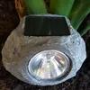 A solar-powered rock-shaped garden light is placed in the soil next to some green plants. Produktbild-Vorschau