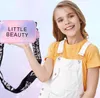 A young girl is holding a small handbag with text on it, and wearing overalls and a t-shirt. Produktbild-Vorschau